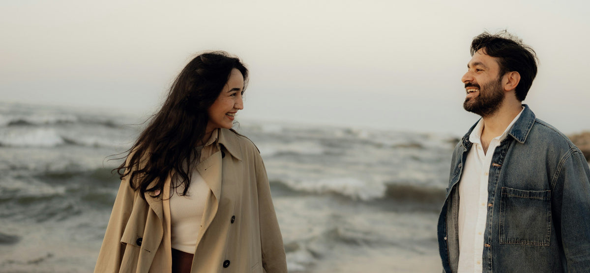 A woman in a trench coat and a man in a denim jacket walking on an autumn beach